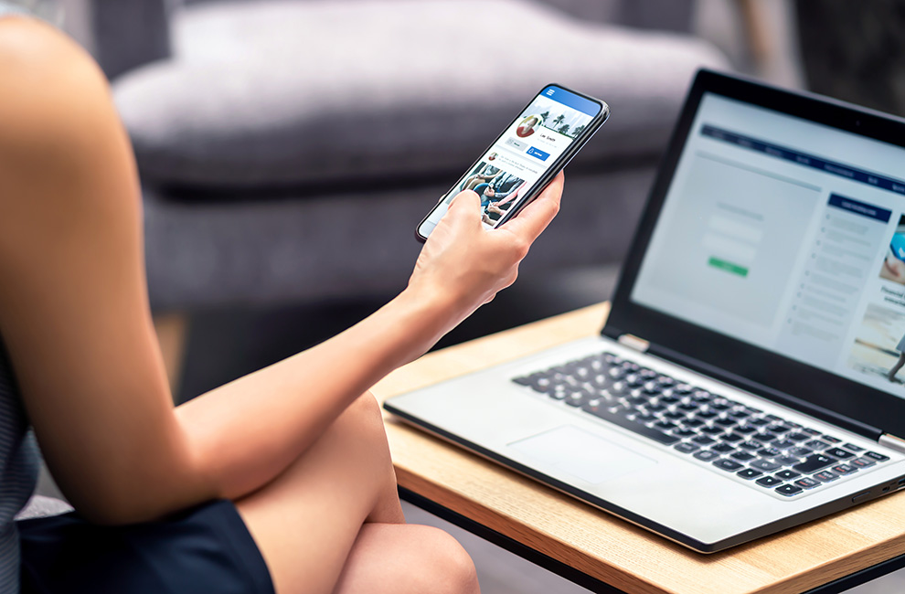 Femme assise devant un ordinateur portable tenant un cellulaire dans sa main ouvert sur l'application Facebook