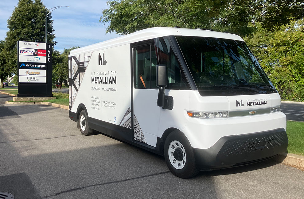 Vue de l'avant et du côté du véhicule de service blanc de Les installations Metalliam avec un wrap partiel avec des photos noir et blanc de structures en acier et une liste des services offerts en-dessous du logo.
