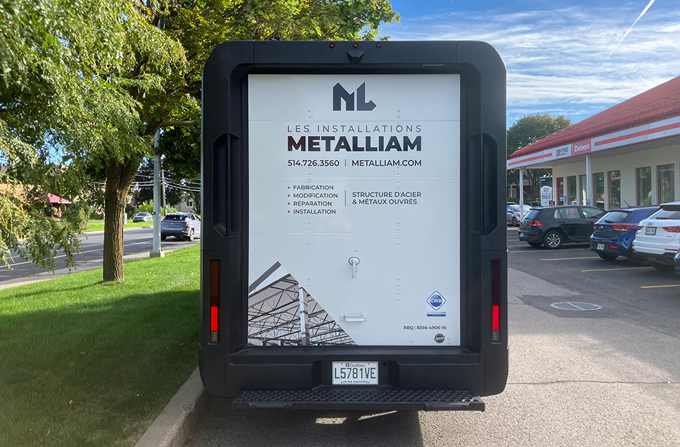 Vue de l'arrière du véhicule de service blanc de Les installations Metalliam avec un wrap partiel avec des photos noir et blanc de structures en acier et une liste des services offerts en-dessous du logo.