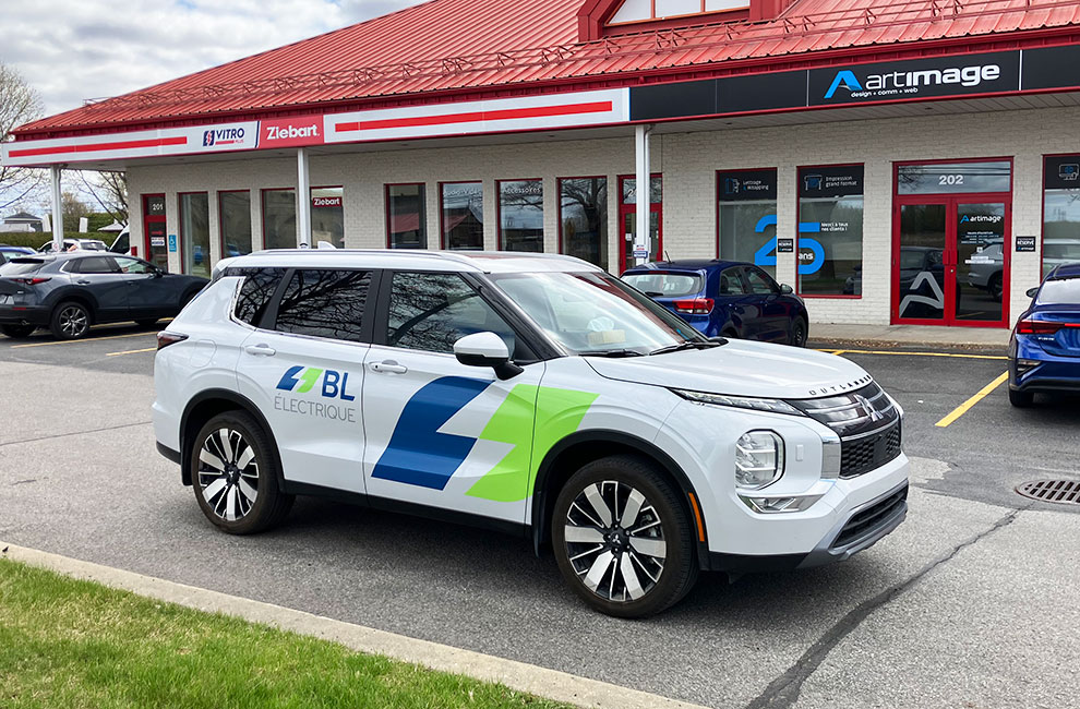 Vue du côté passager du lettrage d'un VUS Mitsubishi Outlander blanc, avec le logo vert, bleu et gris de la compagnie BL Électrique sur les portes avant et arrière