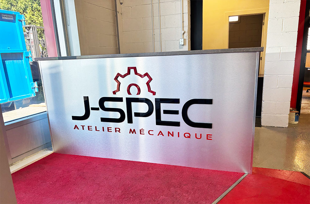 Vue du devant du comptoir d'accueil avec le logo de J-SPEC atelier mécanique en découpe acrylique 3D en noir et rouge sur un panneau d'aluminium brossé