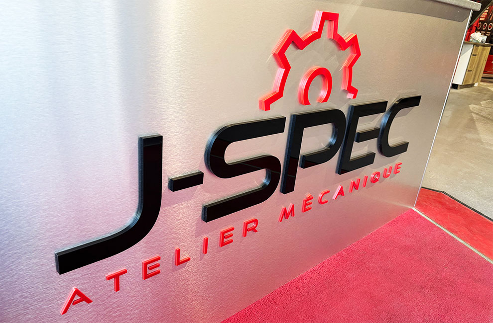 Gros plan sur le logo de J-SPEC atelier mécanique en découpe acrylique 3D en noir et rouge sur un panneau d'aluminium brossé